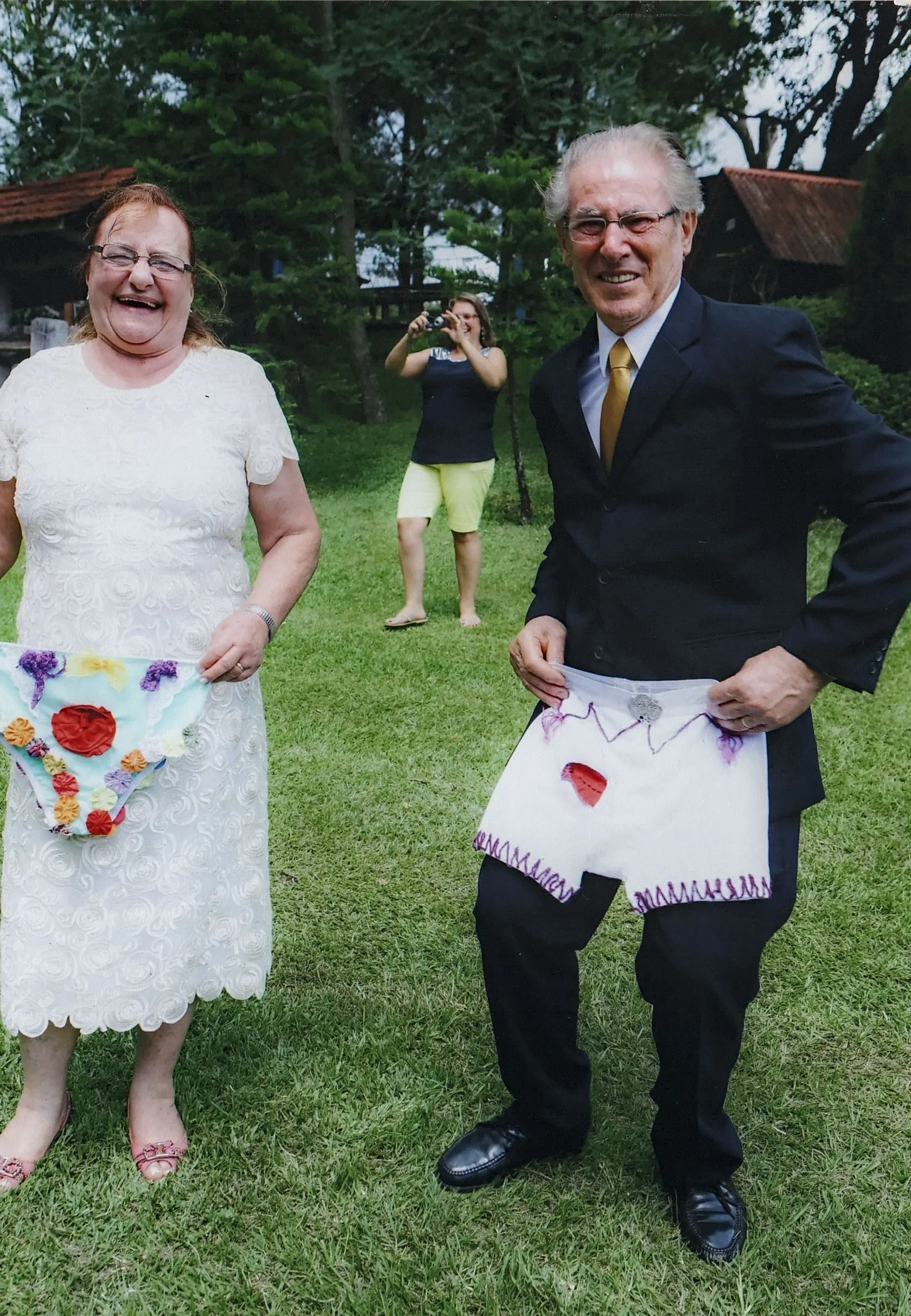 Um homem e uma mulher idosos sorriem enquanto posam em um gramado ao ar livre, segurando aventais com apliques, enquanto uma mulher jovem ao fundo os fotografa.