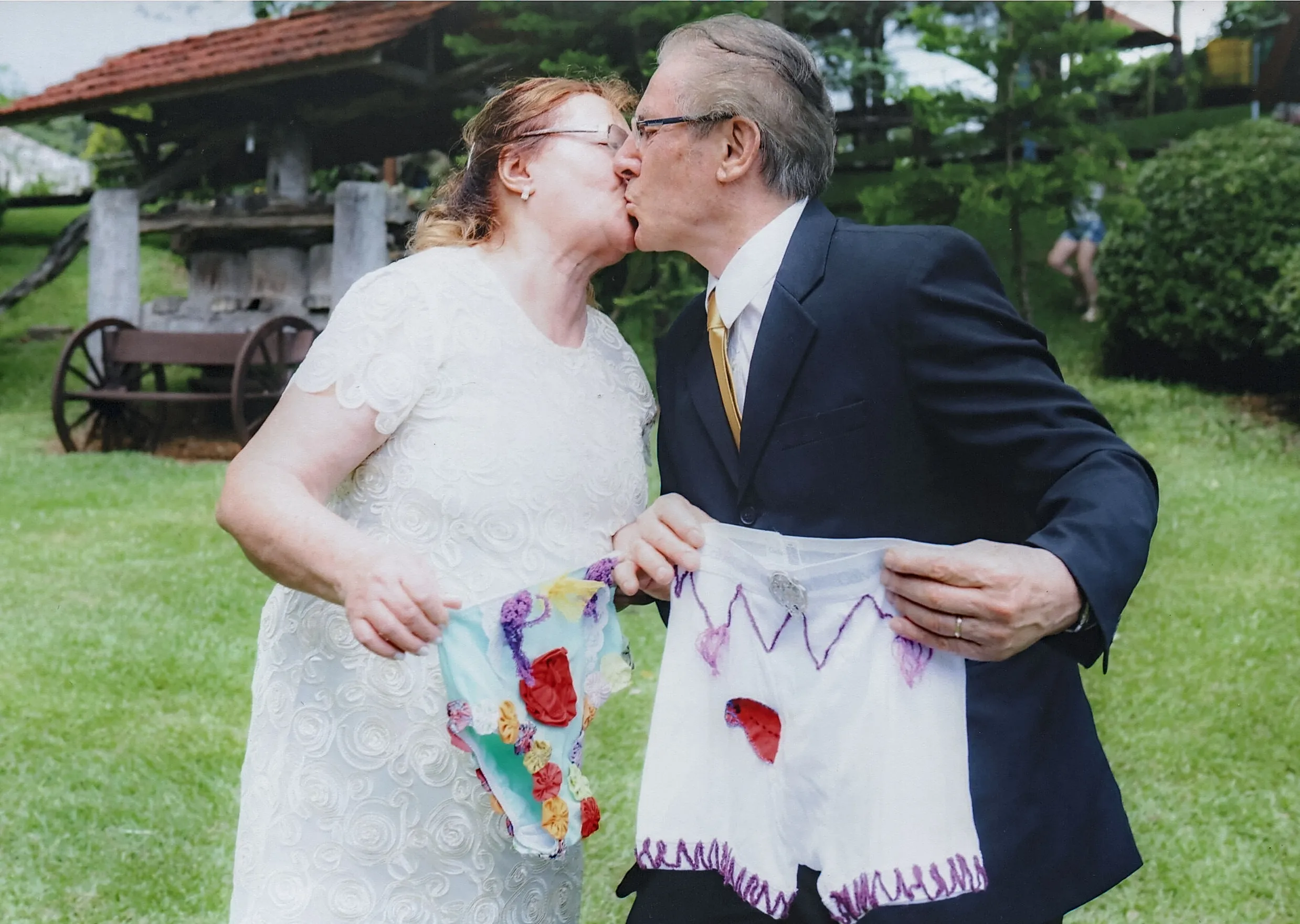 Um casal mais velho se beija enquanto cada um segura uma peça de roupa íntima artesanal de cores vivas em um jardim.