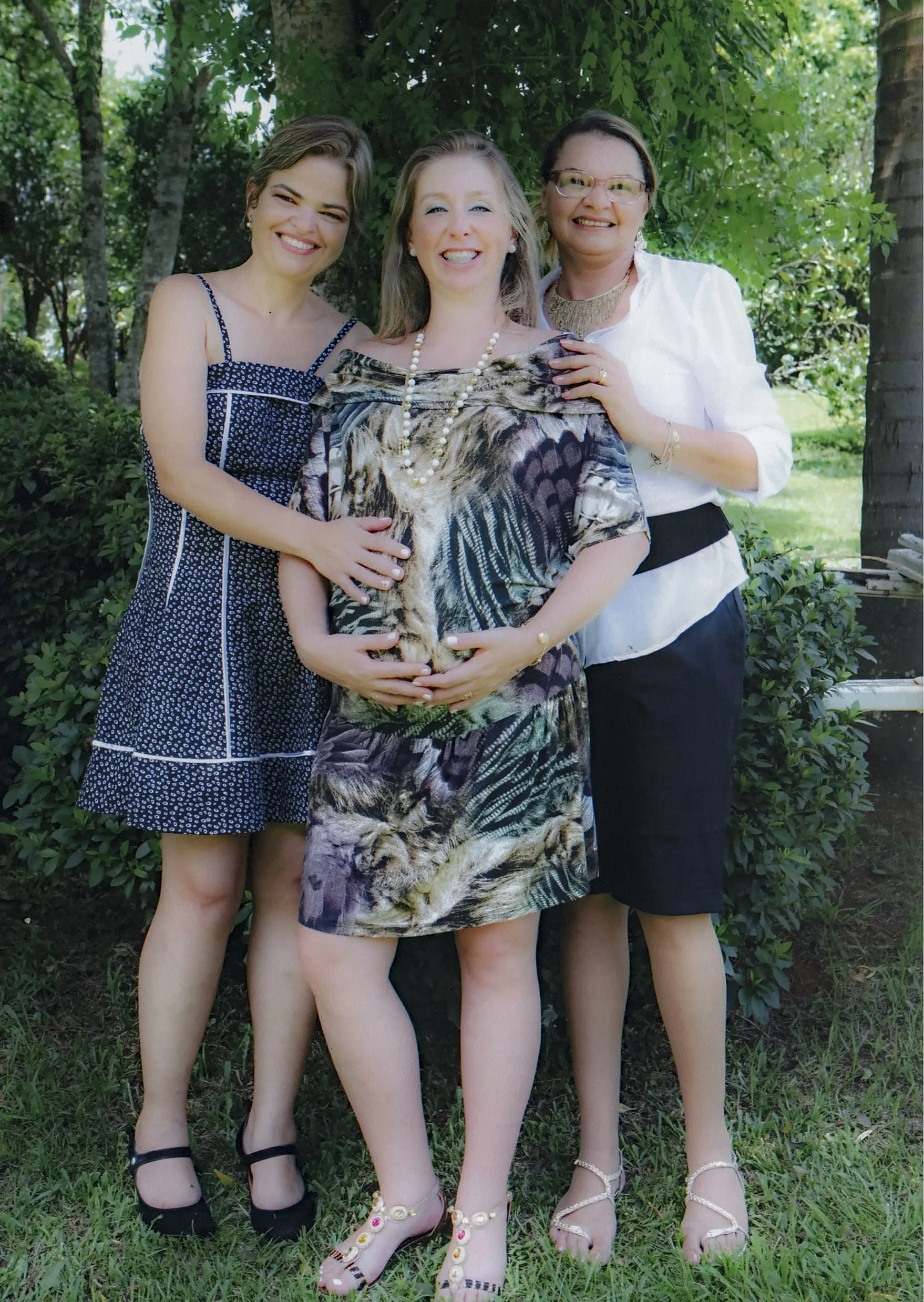 Três mulheres sorridentes posam juntas ao ar livre em um jardim, a mulher do meio está grávida e tem as mãos sobre a barriga, enquanto as outras duas a abraçam.