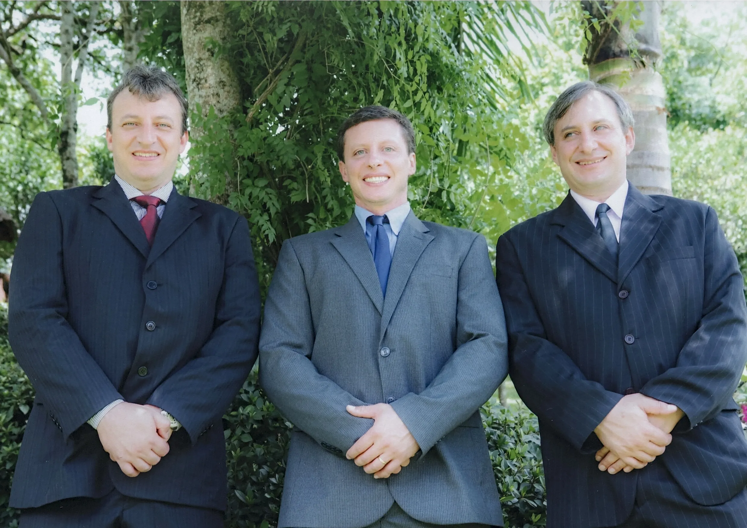 Três homens caucasianos de meia-idade, vestindo ternos escuros, em pé lado a lado na frente de árvores e arbustos verdes, sorrindo para a câmera.