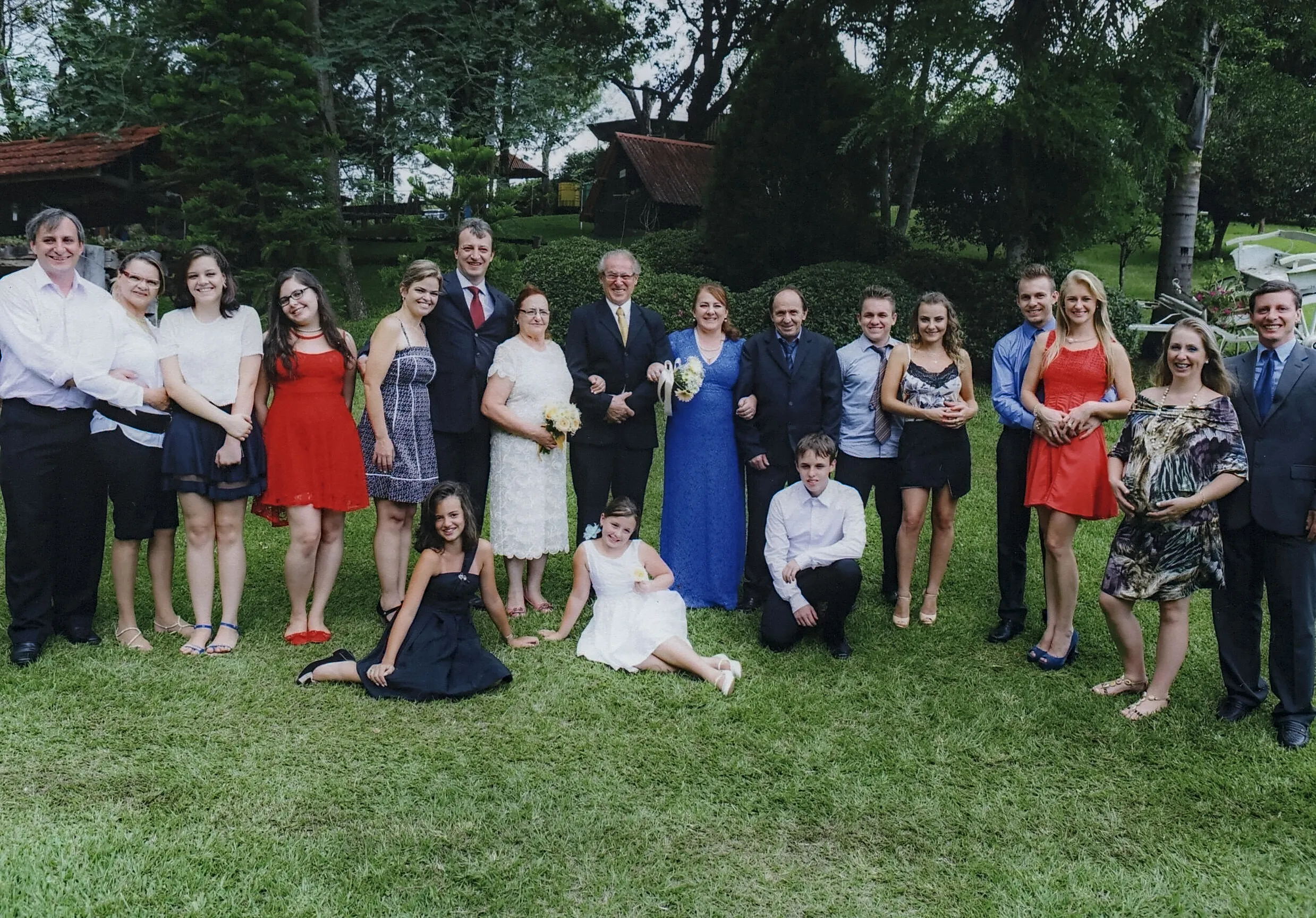 Um grande grupo de pessoas sorridentes, vestidas formalmente, posa para uma foto em um gramado verde com árvores e edifícios ao fundo.