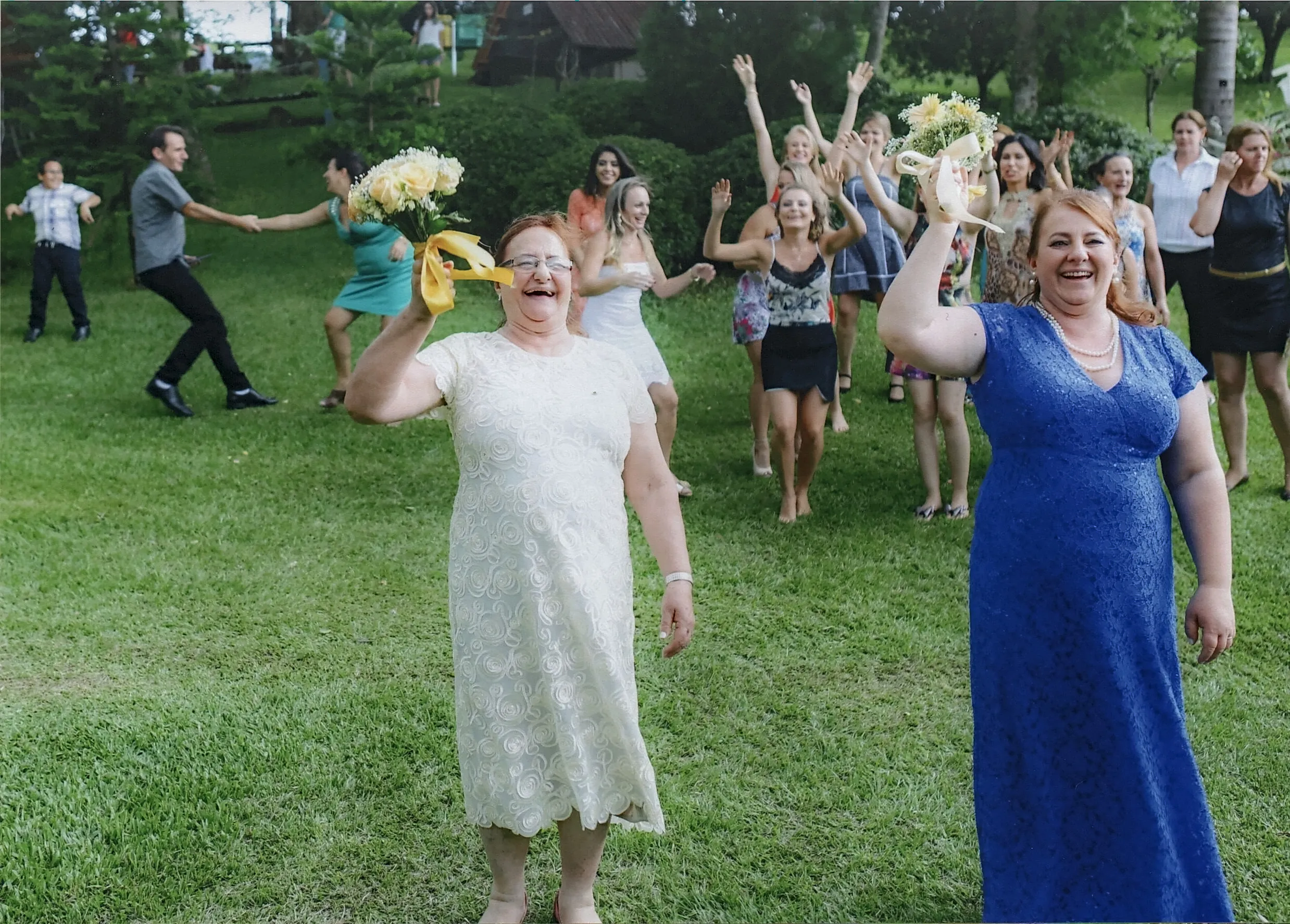 Duas mulheres sorridentes, uma de vestido branco e outra de vestido azul, seguram buquês e estão em primeiro plano. Atrás delas, um grupo de pessoas de diversas idades se diverte em um gramado.
