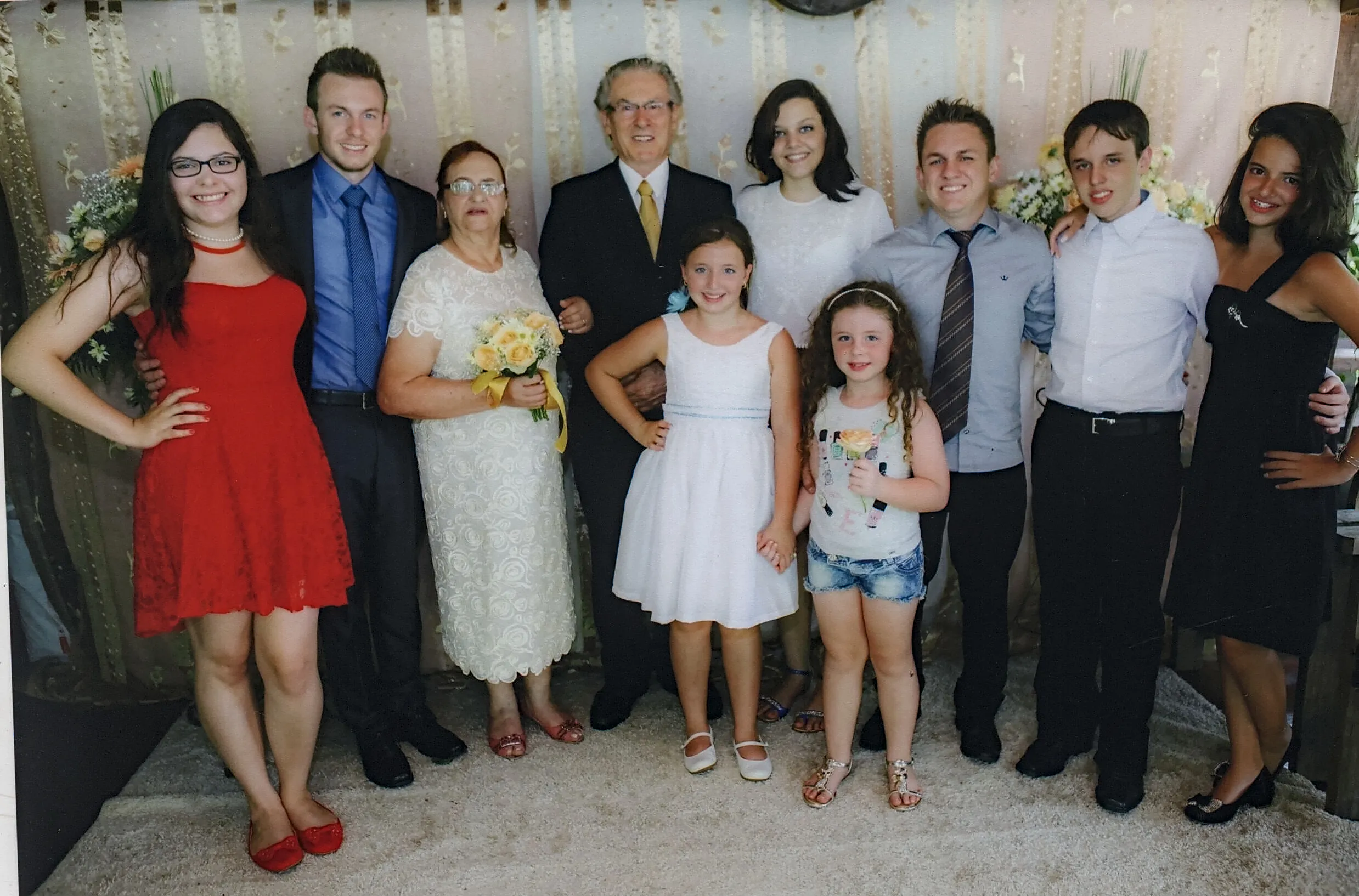 Um grupo de dez pessoas, três homens, cinco mulheres e duas meninas, posa para uma foto de corpo inteiro em uma sala com cortinas bege.