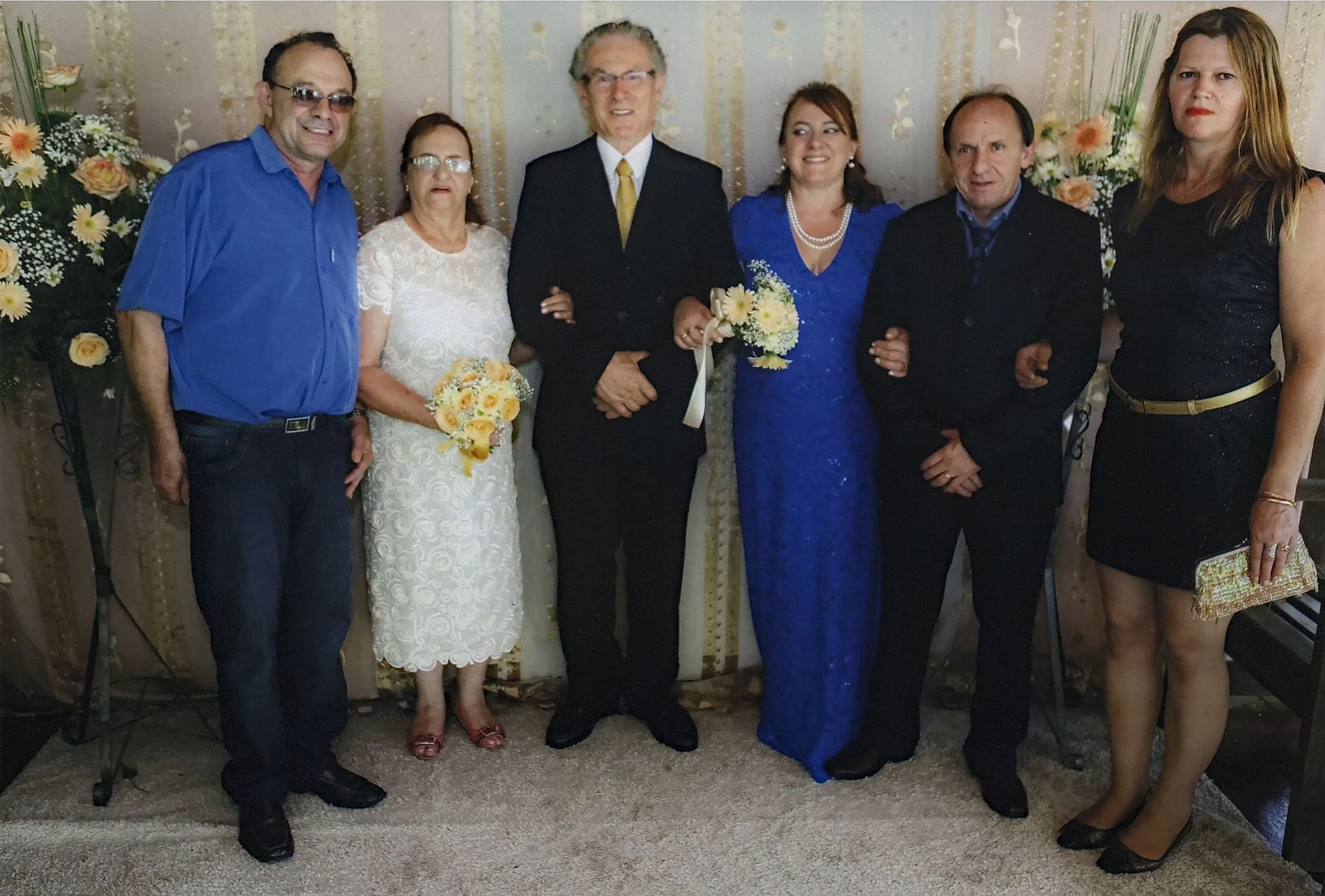 Seis pessoas estão posando para uma foto de grupo, com três homens e três mulheres, todos elegantemente vestidos em um ambiente de celebração, possivelmente um casamento ou evento formal.