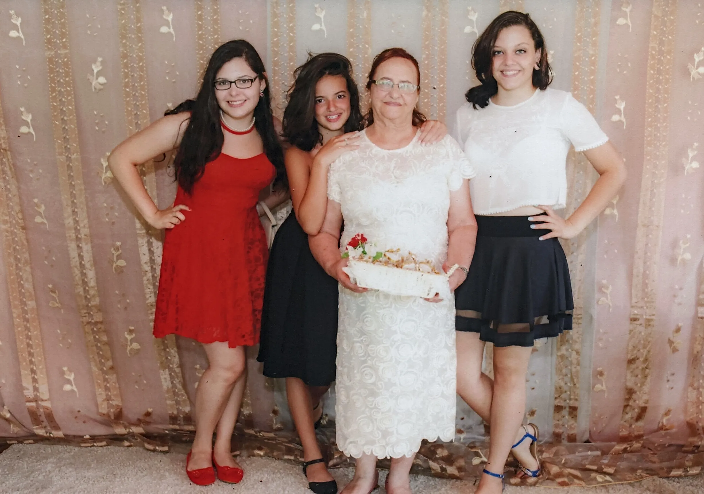 Quatro mulheres, provavelmente de três gerações, posam juntas em uma celebração; a mulher mais velha no centro segura uma cesta decorada, e as outras três a abraçam, todas sorrindo.