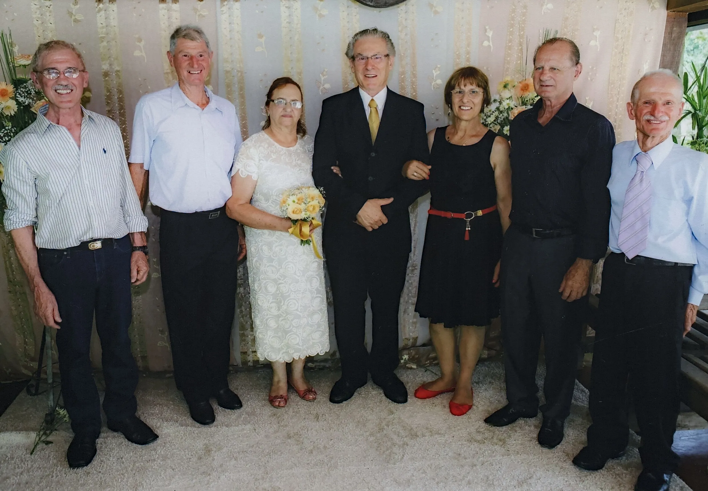 Sete adultos sorridentes, seis homens e uma mulher, posam juntos em frente a uma cortina decorada e um arranjo de flores, provavelmente em um casamento ou evento formal.