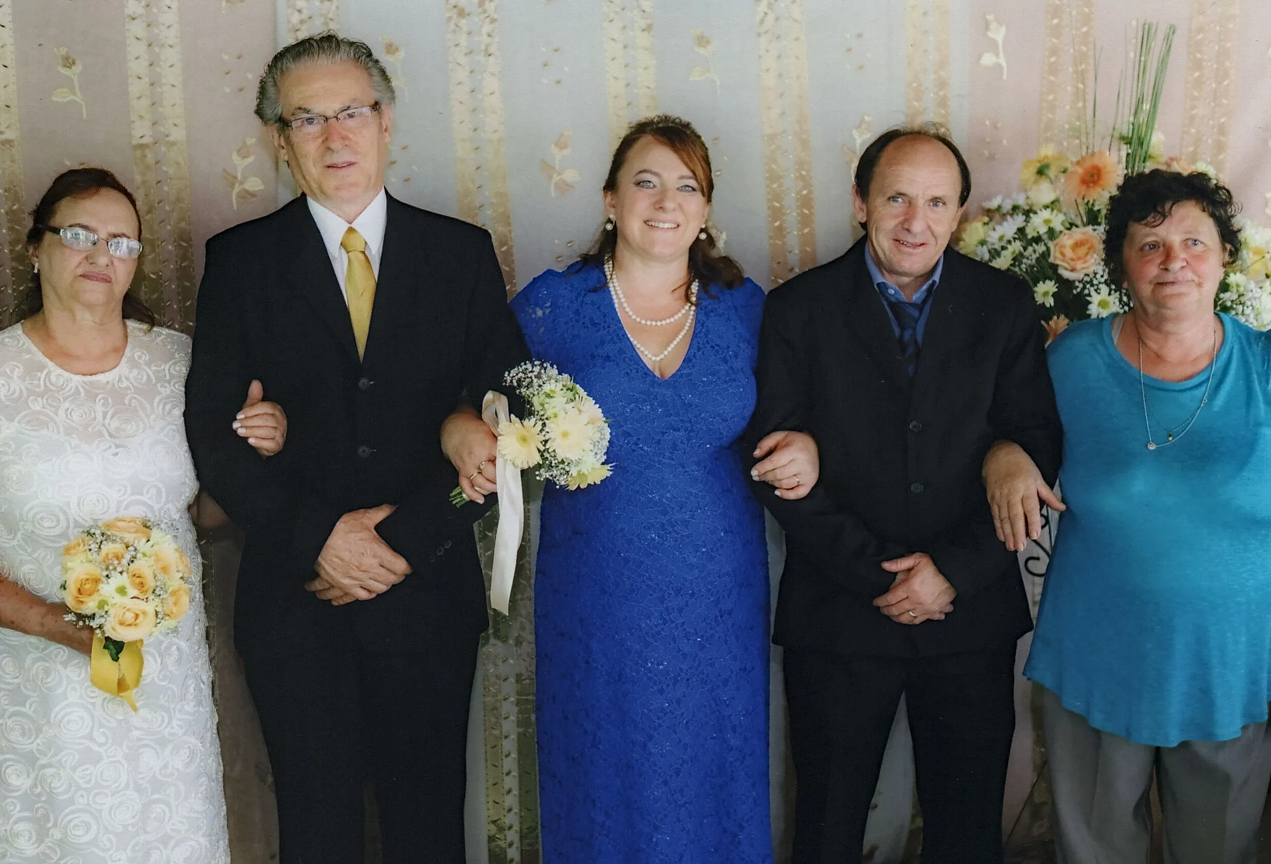 Cinco pessoas estão em pé em uma fileira horizontal contra uma parede estampada em tons de creme. Da esquerda para a direita: uma mulher mais velha de vestido branco com um buquê, um homem de terno preto e gravata dourada, uma mulher de vestido azul vibrante com um buquê e um colar de pérolas, um homem de terno preto e uma mulher de blusa azul e calça cinza. As pessoas de terno estão de braços dados com as mulheres ao lado delas.