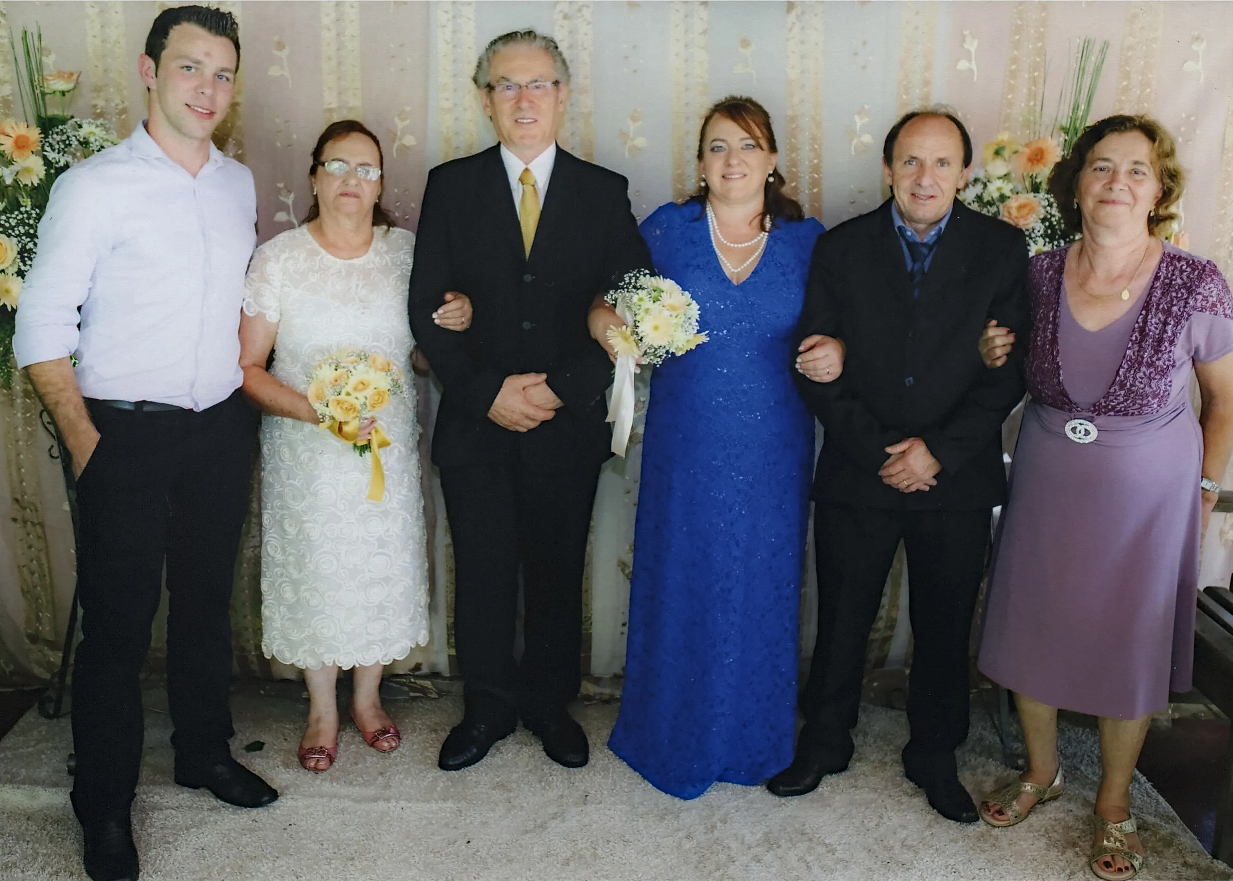 Seis pessoas, incluindo um homem de camisa branca, um homem e uma mulher em trajes de casamento brancos e azuis, e outros casais em roupas formais, posam para uma foto em um evento formal.