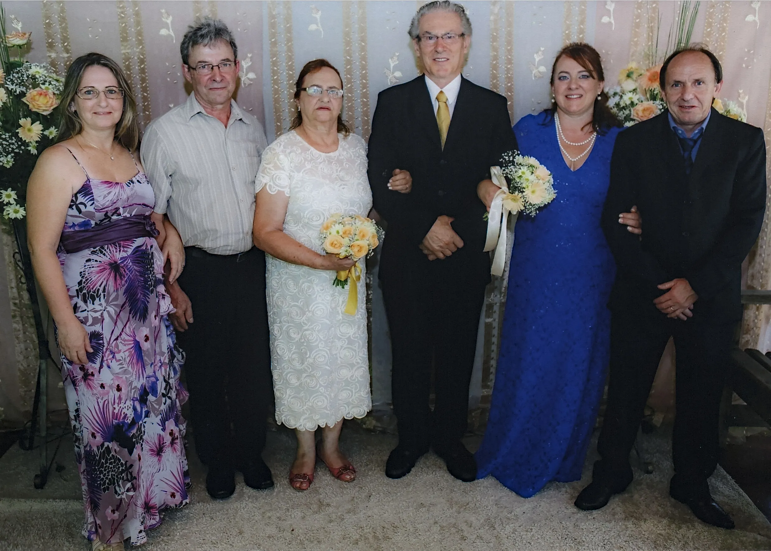 Seis pessoas sorridentes em um casamento. A noiva está em um vestido branco e segura um buquê de flores amarelas. O noivo está de terno preto e gravata dourada. A mulher à esquerda da noiva está de vestido floral roxo, e o homem à esquerda dela está de camisa xadrez. A mulher à direita do noivo está de vestido azul, e o homem à direita dela está de terno preto.