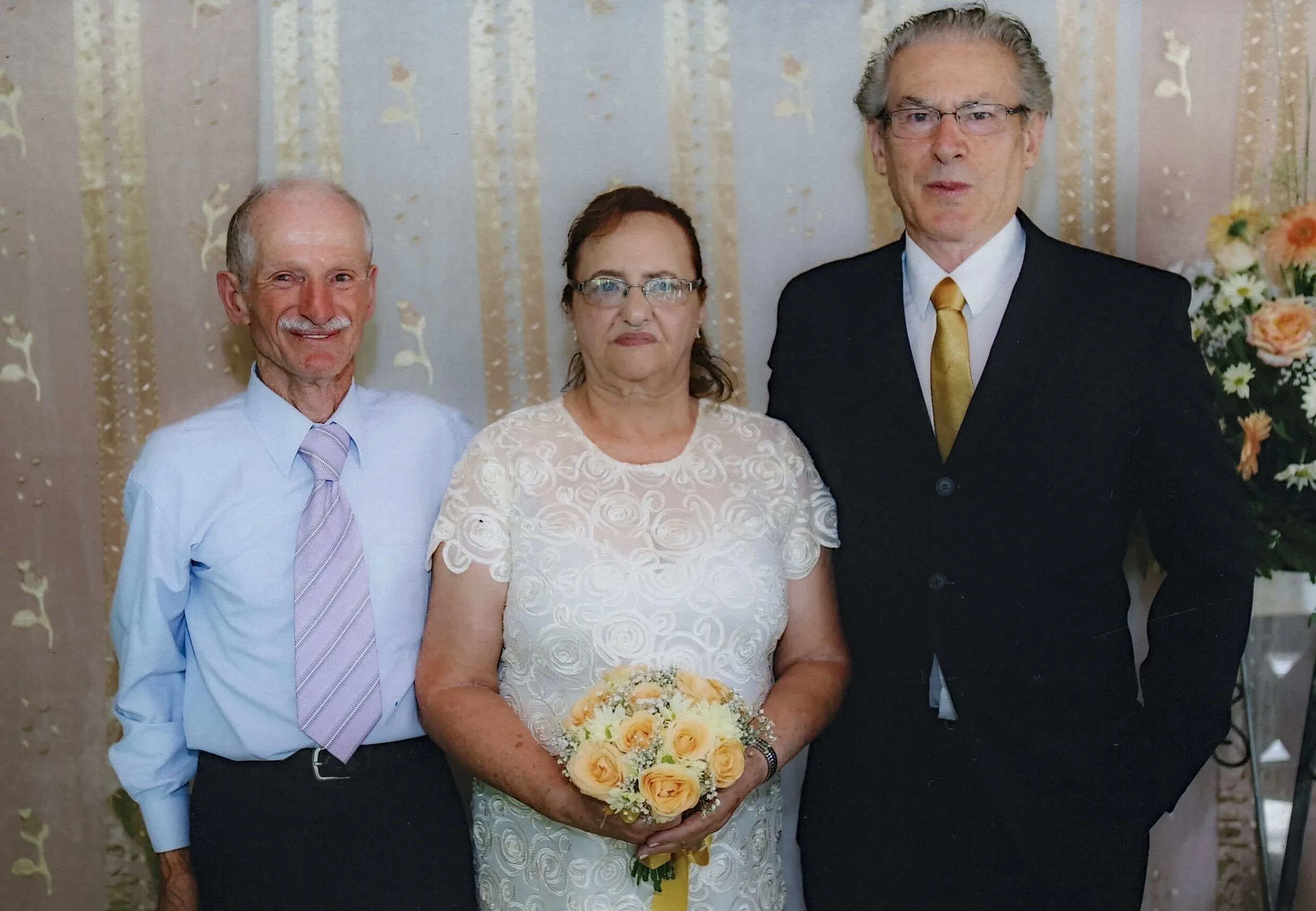 Três pessoas adultas em trajes de casamento posam na frente de uma parede com cortinas claras; a noiva no centro usa um vestido branco de renda e segura um buquê de rosas pêssego, ladeada por dois homens de terno e gravata.