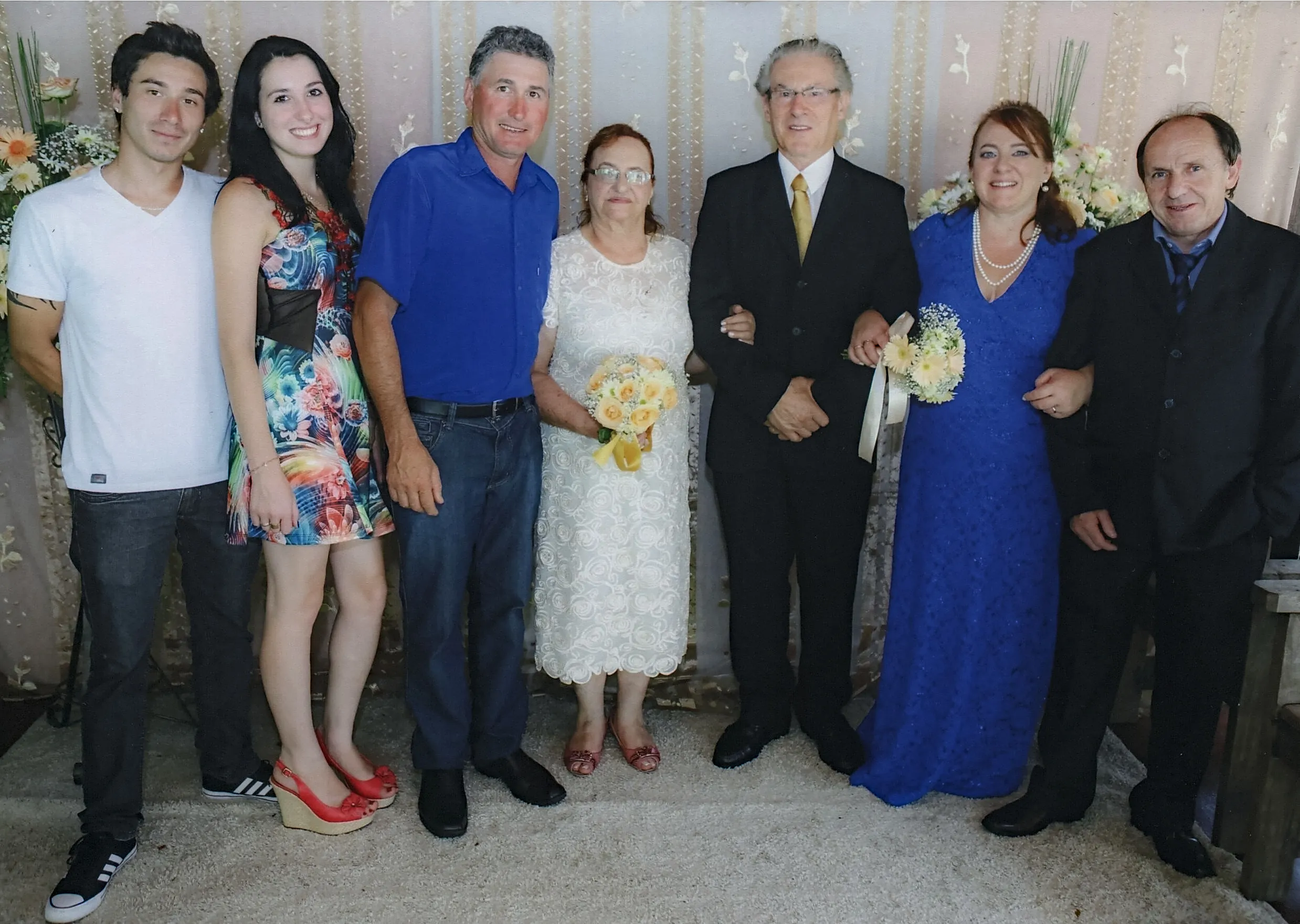 Sete pessoas, incluindo noivos com buquês, posam lado a lado em um evento formal, possivelmente um casamento.