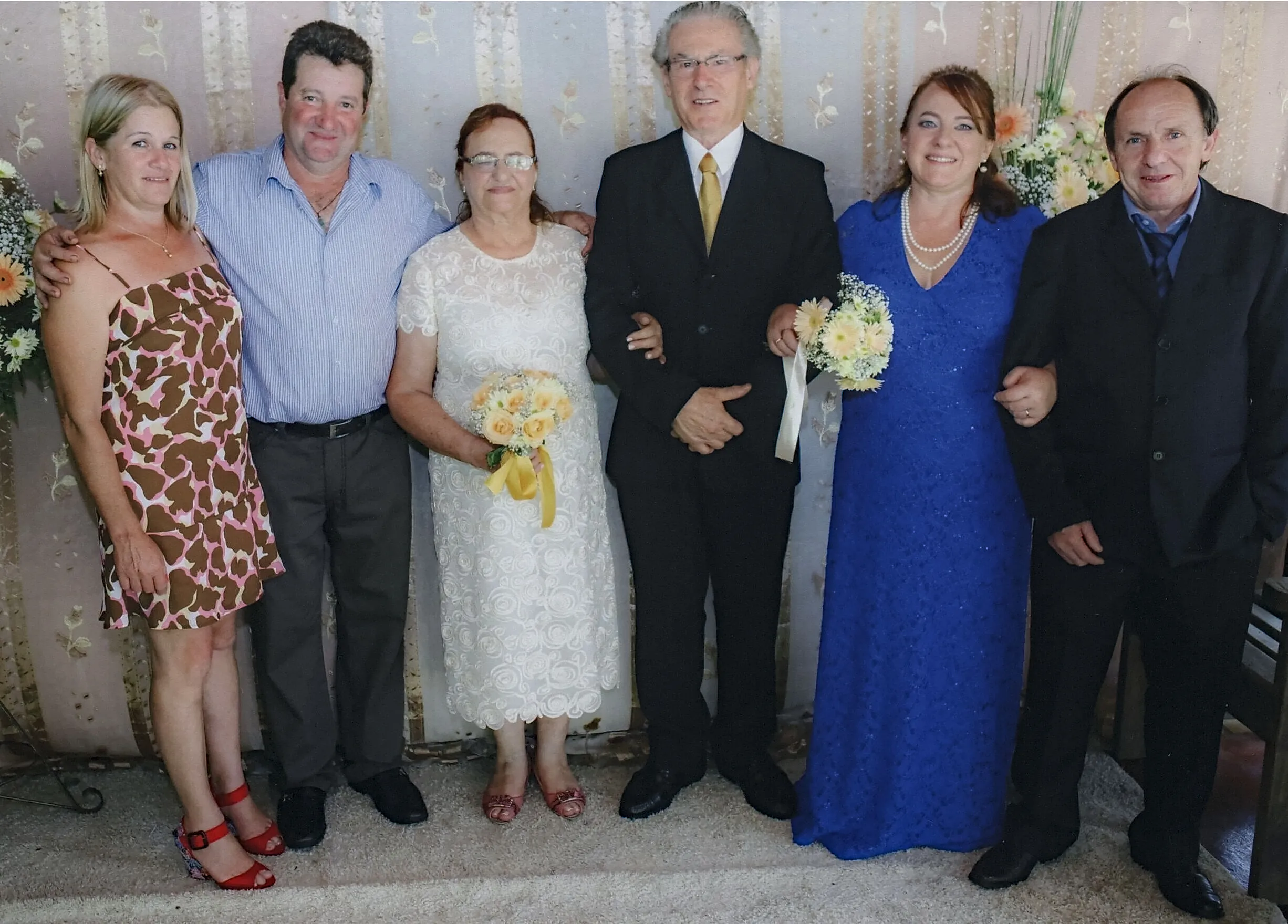 Seis adultos sorrindo, três homens e três mulheres, em trajes formais, posam para uma foto de grupo, com um buquê de flores amarelas sendo segurado por uma mulher de vestido branco, e outro buquê nas mãos da mulher de vestido azul.