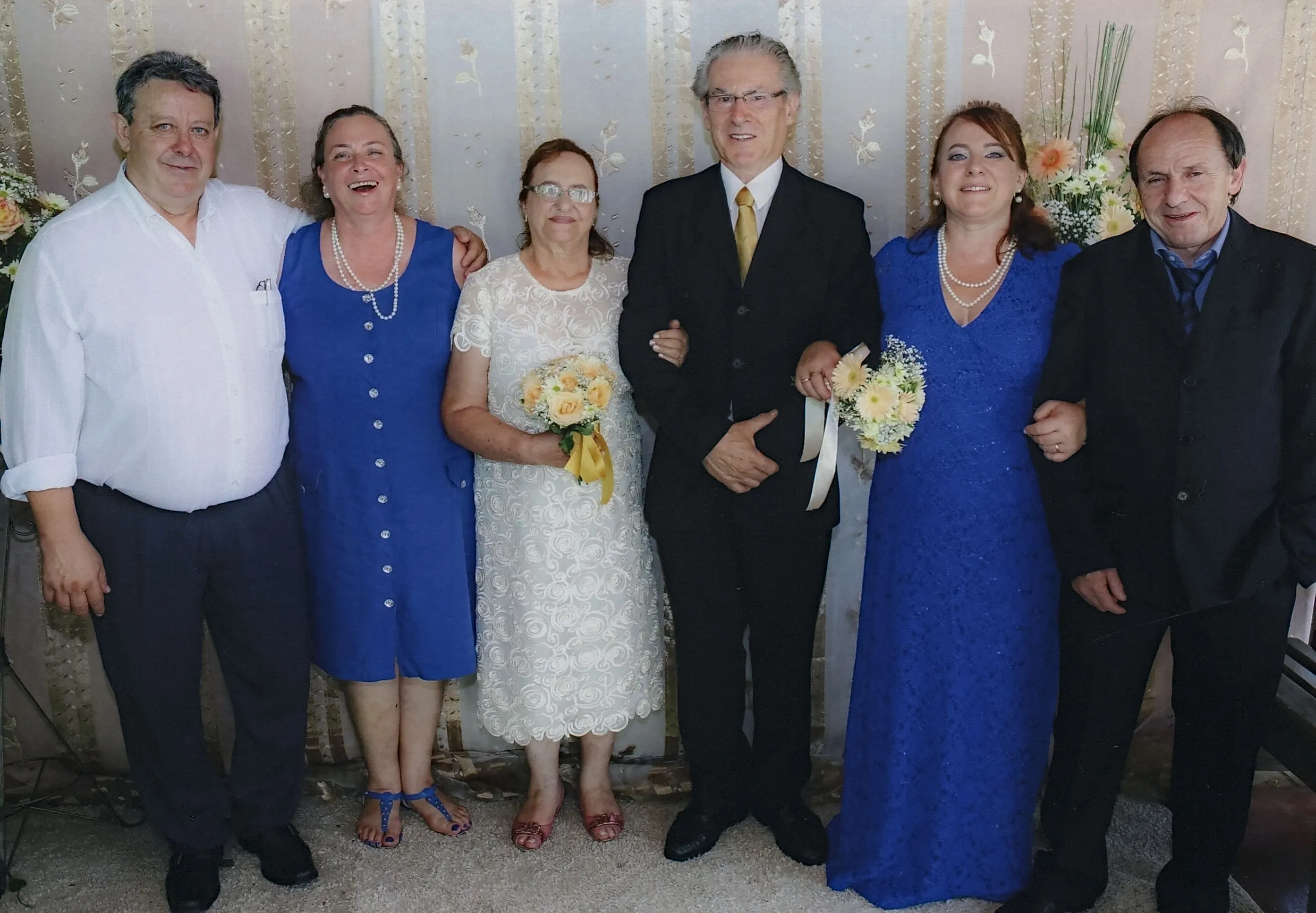 Seis adultos estão em pé, lado a lado, em uma linha, dentro de uma sala. O homem do meio veste um terno preto com uma camisa branca e uma gravata amarela. À esquerda dele, uma mulher de vestido branco segura um buquê de flores amarelas. À esquerda dela, uma mulher de vestido azul. À esquerda dela, um homem de camisa branca e calças escuras. À direita do homem do meio, uma mulher de vestido azul, segurando um buquê de flores amarelas e brancas, e um homem de terno preto.