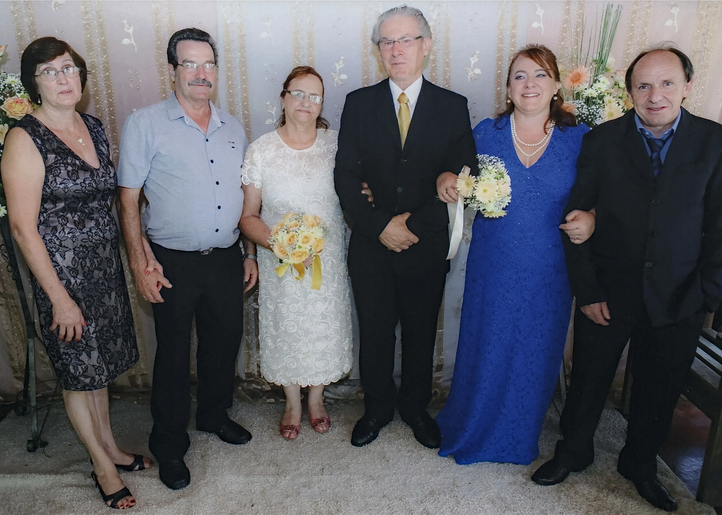 Seis adultos de pé em um evento formal, três homens e três mulheres, todos olhando para a câmera, com flores e cortinas no fundo.