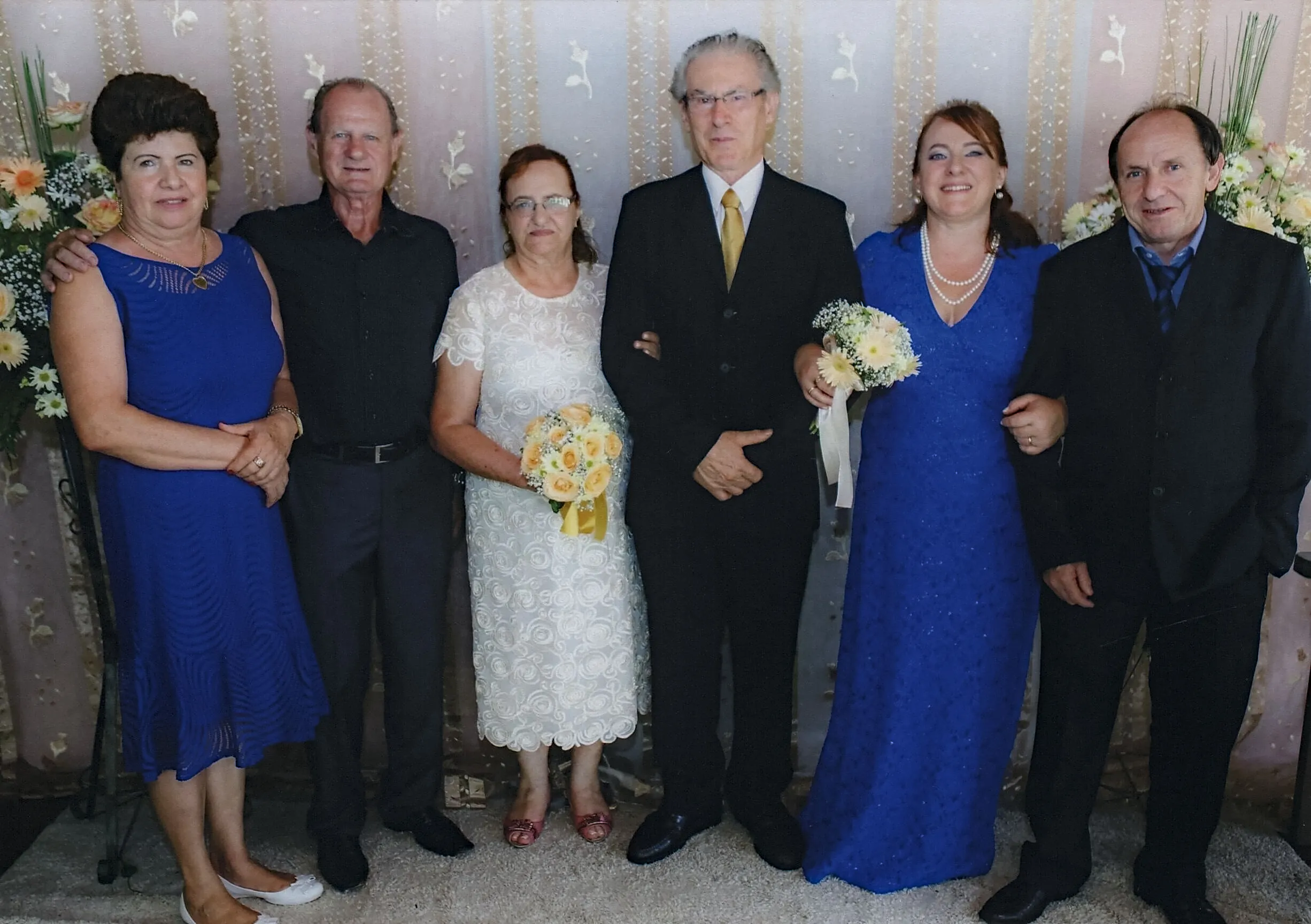 Três casais adultos posando para uma foto de família em um evento, com a noiva no centro em um vestido branco e os outros em roupas formais escuras e azuis.