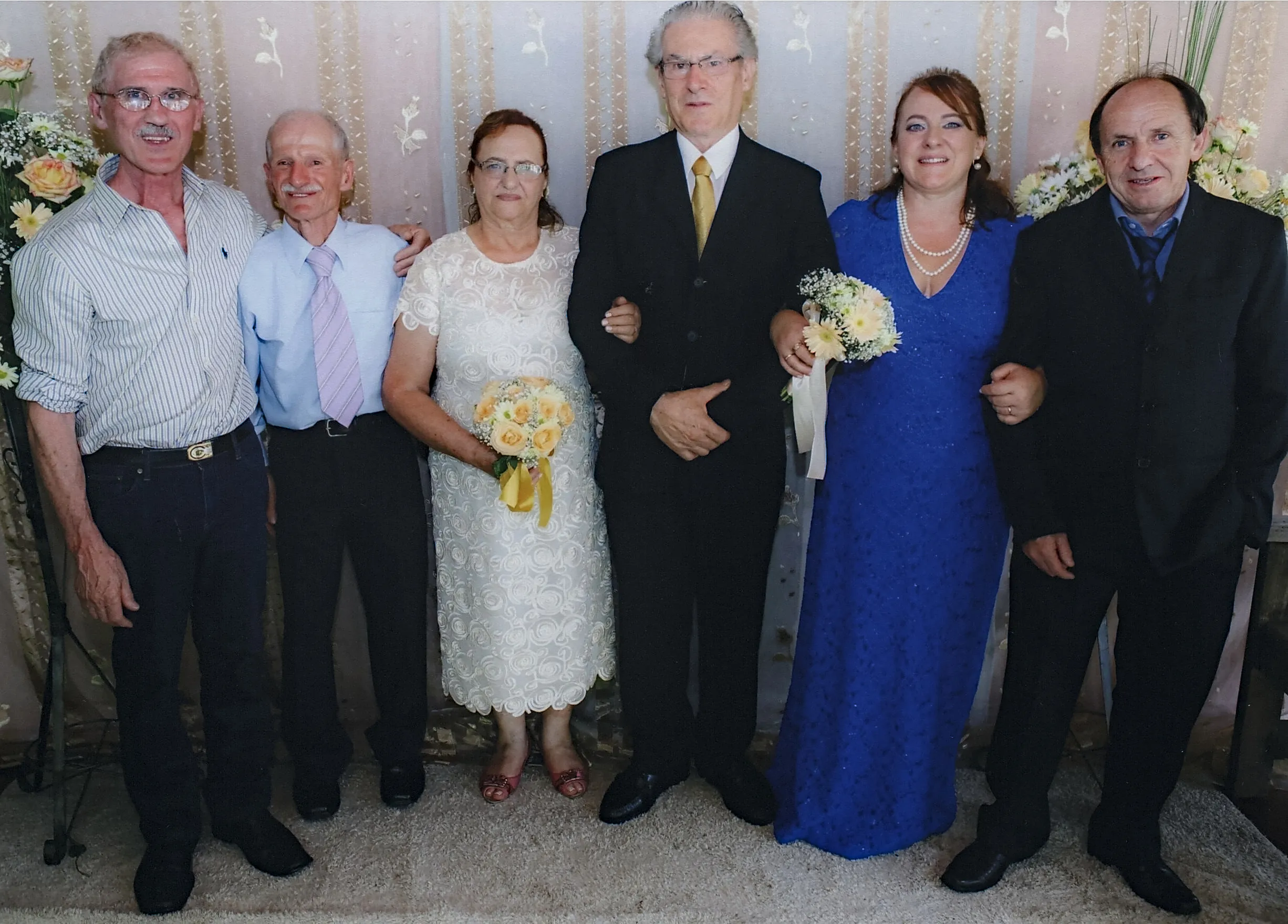 Seis pessoas em trajes formais em um casamento: três homens em ternos escuros, duas mulheres em vestidos claros segurando buquês e um homem de camisa listrada e jeans, todos em pé dentro de casa.