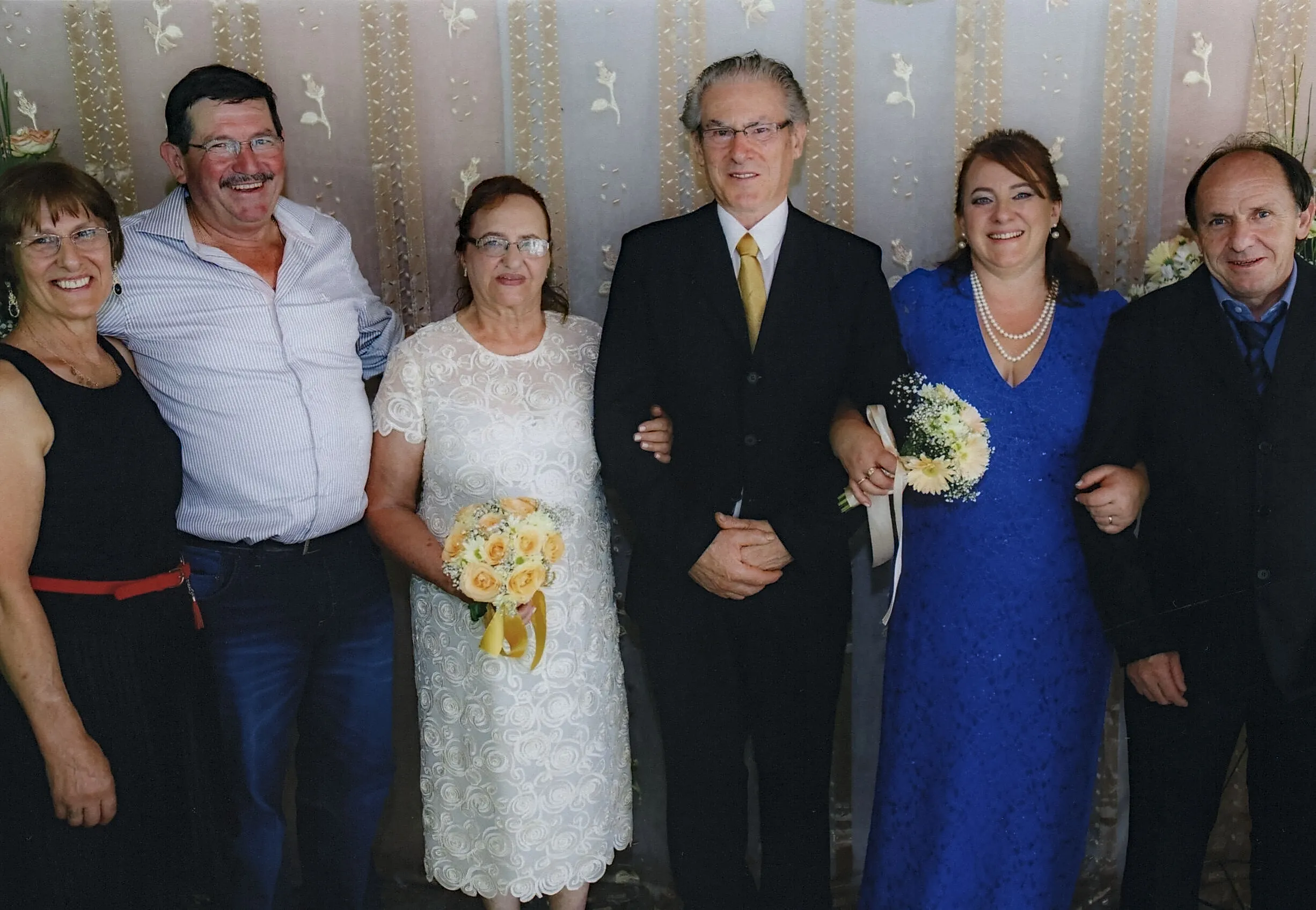 Seis adultos posam para uma foto em um evento formal, com três casais lado a lado; o casal do meio é o foco principal.