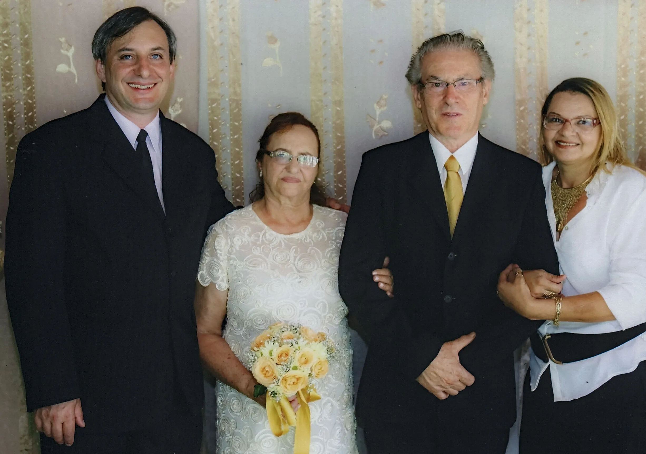 Um homem em um terno preto com uma camisa rosa e gravata escura, uma mulher em um vestido de noiva branco segurando um buquê de rosas amarelas, um homem mais velho em um terno preto com uma camisa branca e gravata amarela, e uma mulher em uma camisa branca com uma gola dourada e óculos em um evento formal.