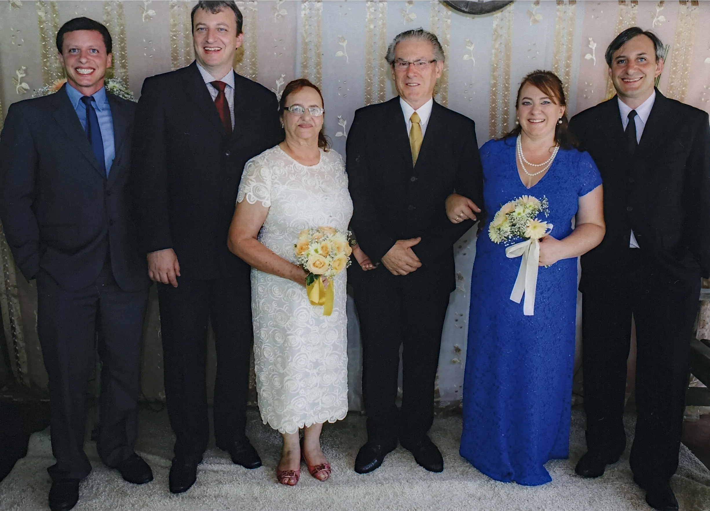 Seis adultos, três homens e três mulheres, estão em fila para uma foto, em um quarto com cortinas bege e uma parede clara com padrões dourados. A mulher do meio veste um vestido branco e segura um buquê de noiva. O homem ao seu lado veste um terno preto com uma camisa branca e gravata amarela. Os outros quatro estão em trajes formais.