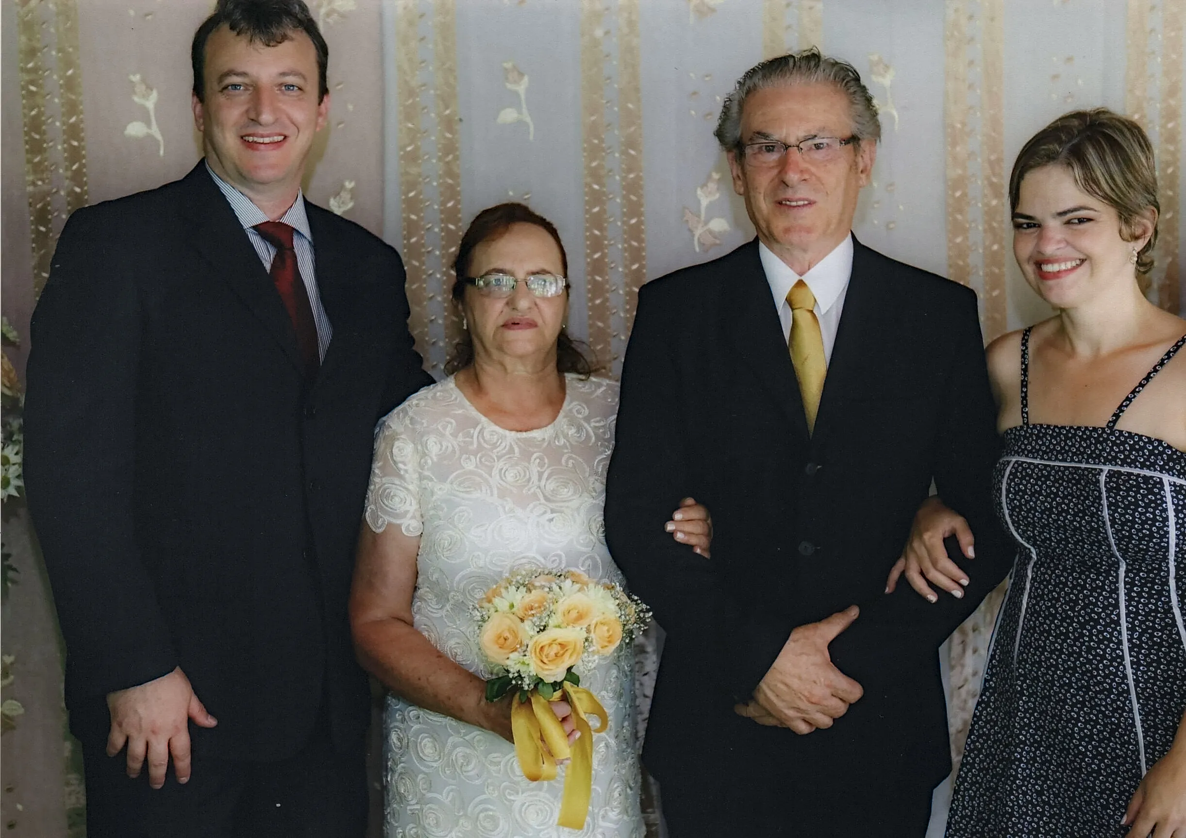Quatro pessoas em roupas formais posam para uma foto em um casamento, com uma mulher de vestido branco no centro segurando um buquê e uma mulher de vestido preto à direita.