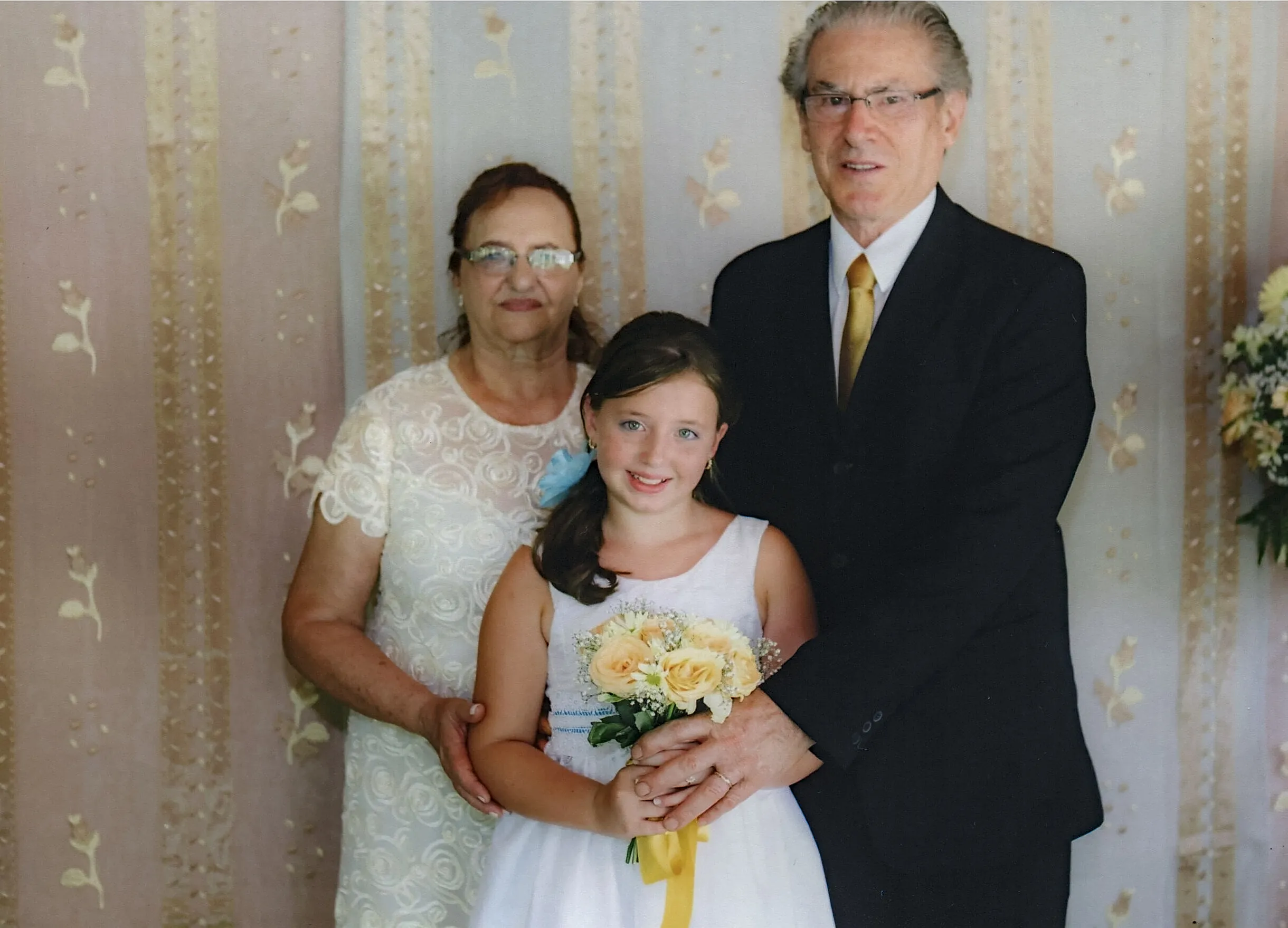 Menina sorridente segurando buquê de rosas amarelas entre um homem e uma mulher mais velhos, que sorriem gentilmente.