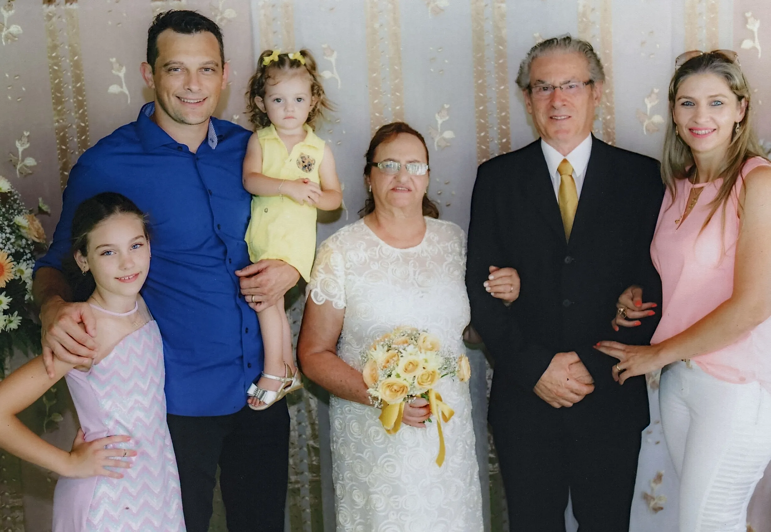 Grupo de seis pessoas, incluindo duas crianças, em pé e posando para uma foto em um cenário claro e decorado, provavelmente em um evento formal.