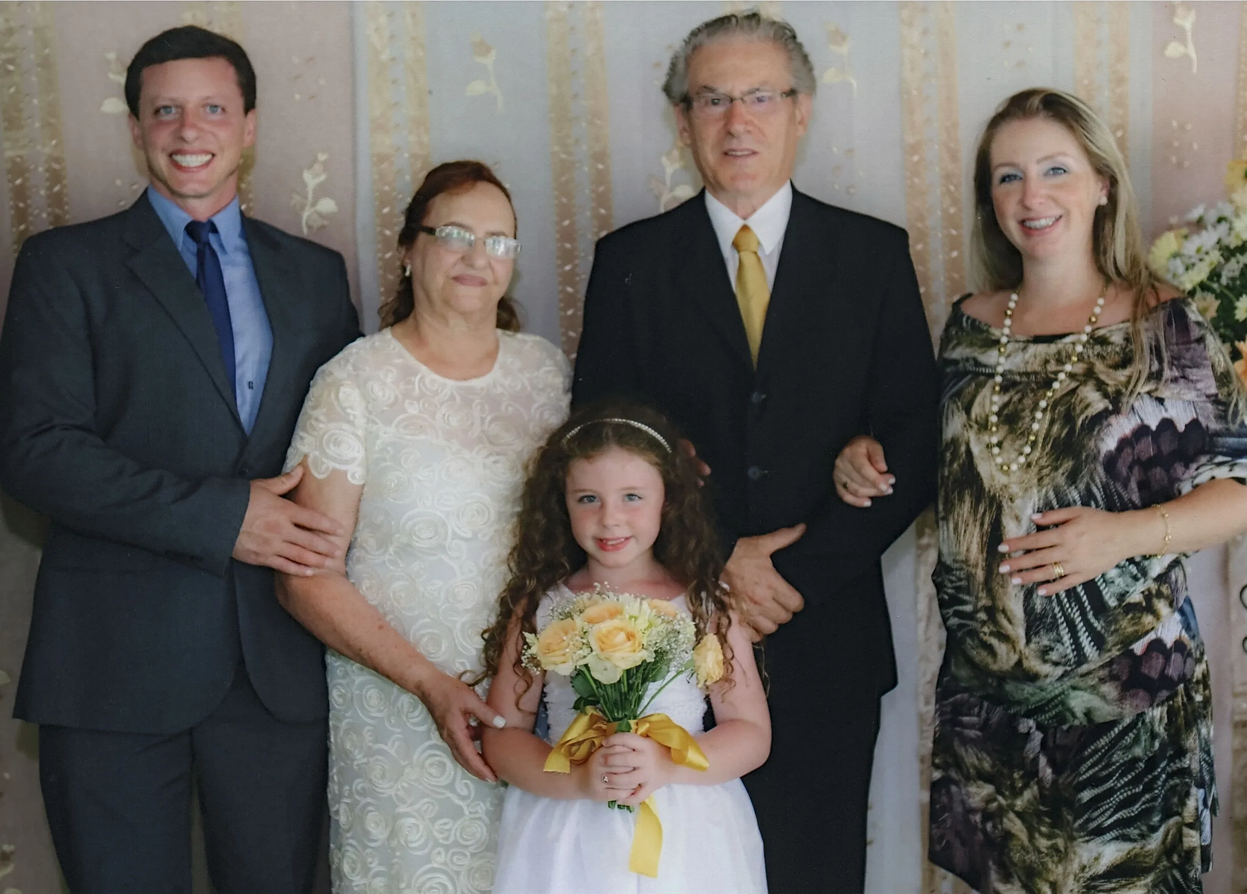 Um homem de terno, uma mulher de vestido branco, uma menina de vestido branco segurando um buquê, um homem de terno e uma mulher de vestido estampado, todos posando juntos.