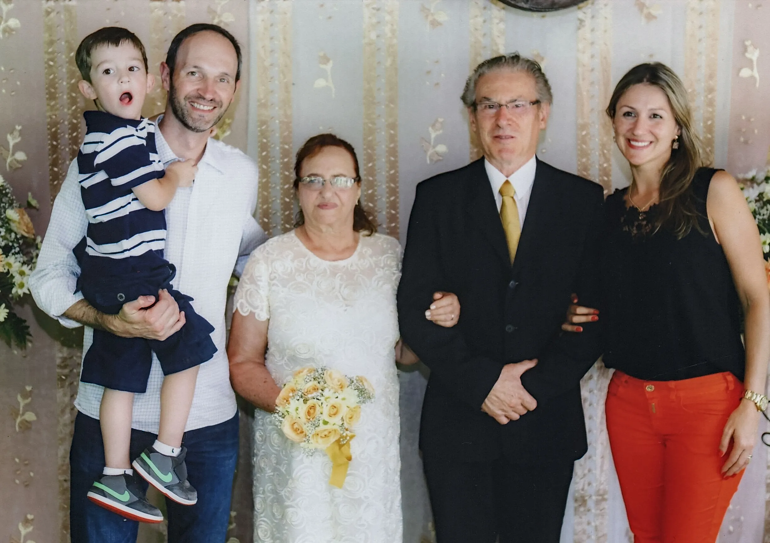 Um menino pequeno de camisa polo listrada e shorts escuros está no colo de um homem barbudo de camisa branca e jeans, enquanto uma mulher de vestido branco, segurando um buquê de flores amarelas e brancas, e um homem de terno preto e gravata dourada, estão parados ao lado de uma mulher sorridente de blusa preta e calças vermelhas.