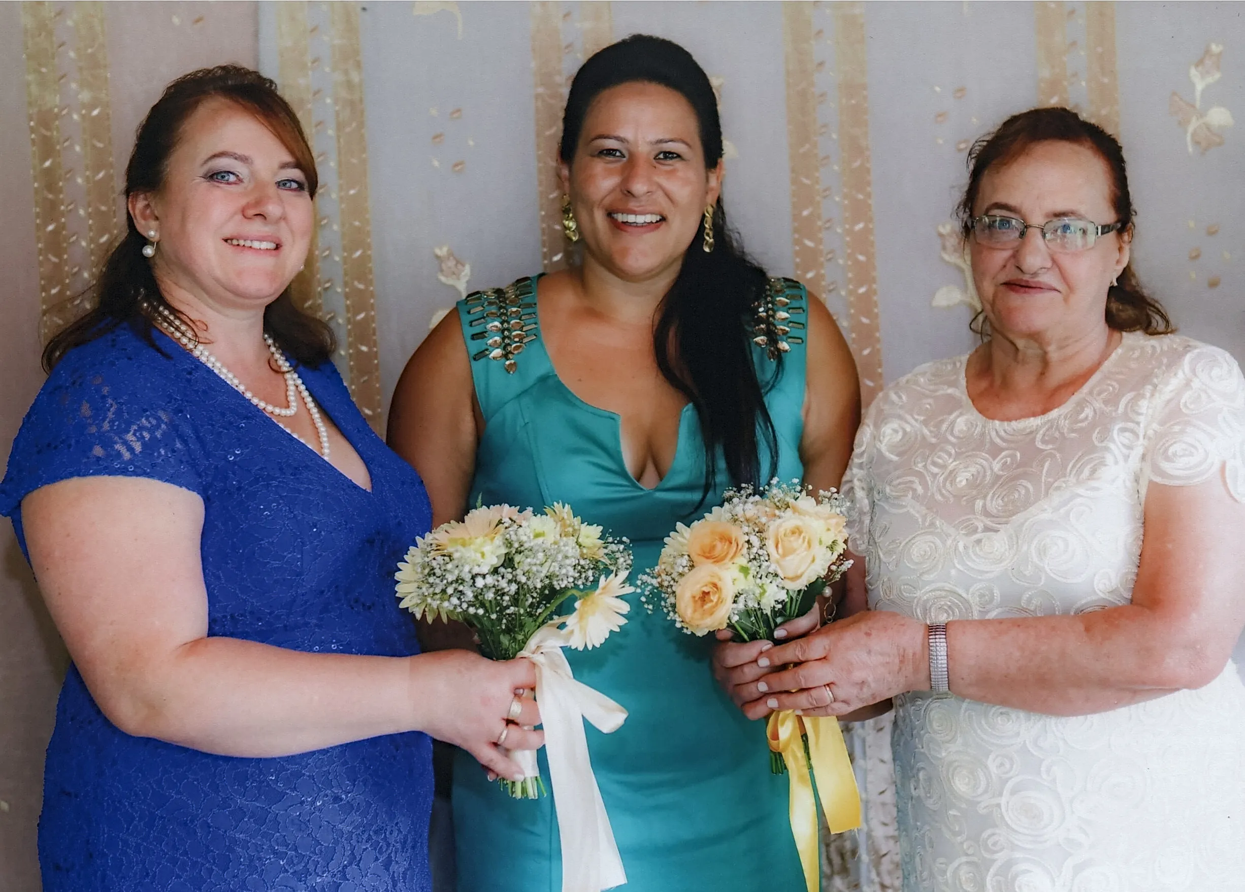 Três mulheres sorrindo para a câmera, segurando buquês de flores; a da esquerda usa um vestido azul de renda e um colar de pérolas, a do meio um vestido turquesa com detalhes dourados nos ombros, e a da direita um vestido branco de renda e óculos.