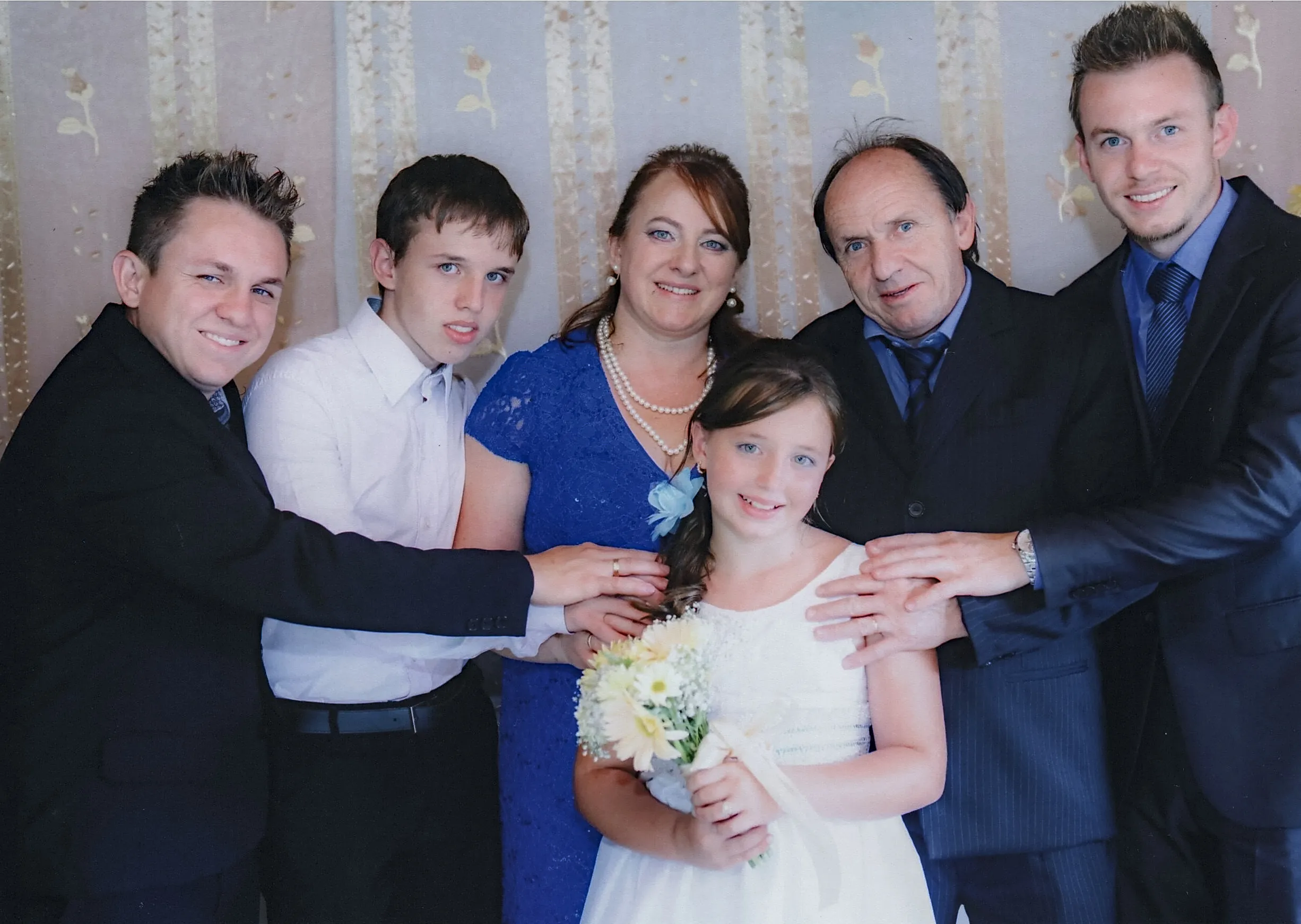 Um grupo de seis pessoas, incluindo uma jovem vestida de branco segurando um buquê, posa para uma foto em um evento formal, com as mãos de vários membros da família descansando uns sobre os outros em apoio.