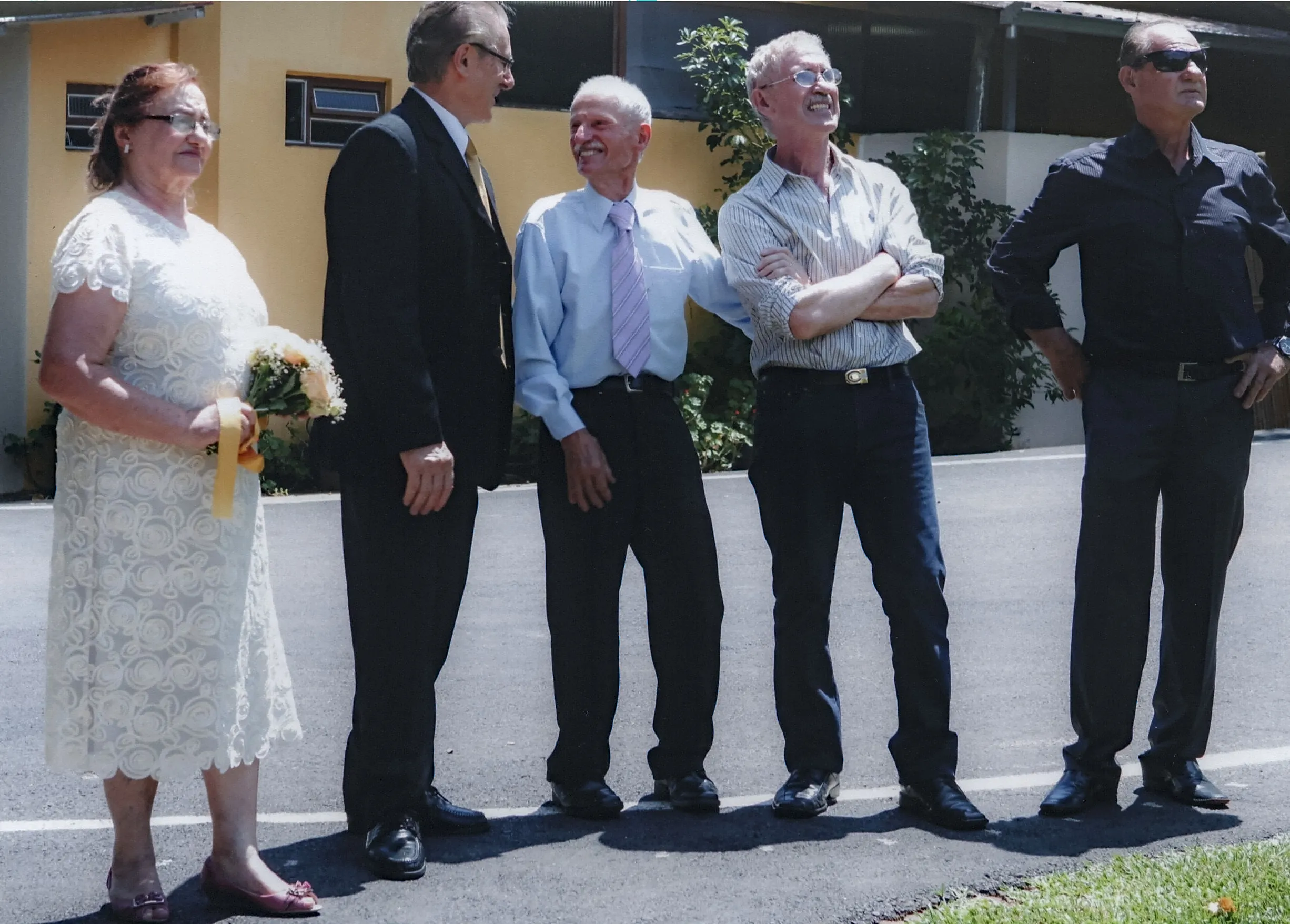Cinco indivíduos, quatro homens e uma mulher, em pé em uma calçada de asfalto perto de um prédio amarelo e folhagem verde.