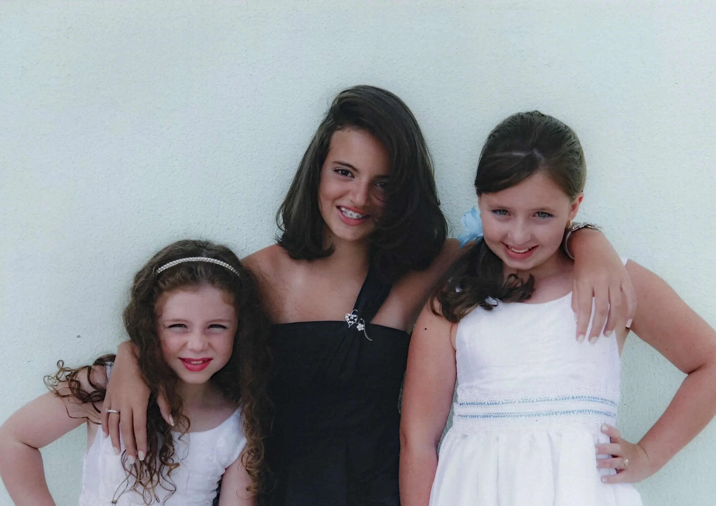 Três meninas, a do meio mais alta, sorrindo para a câmera, com os braços uma da outra, em frente a uma parede lisa.