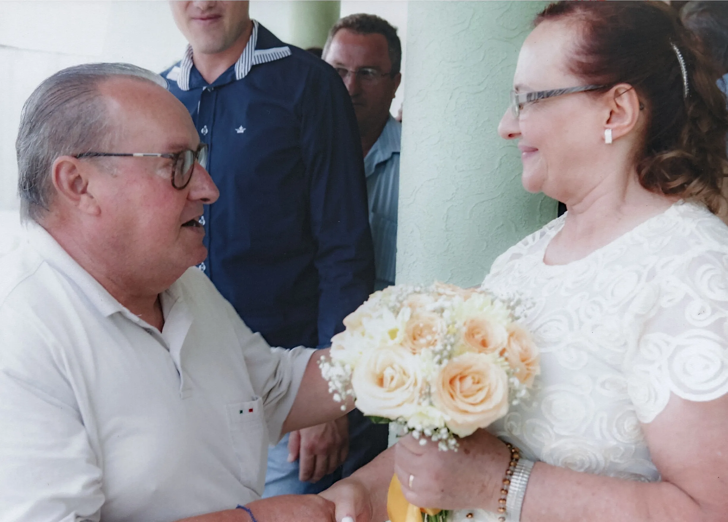 Um homem mais velho com óculos e camisa polo bege sorri para uma mulher de óculos com um vestido de renda branca, que segura um buquê de rosas pêssego e brancas, enquanto se cumprimentam com as mãos, com duas figuras masculinas desfocadas ao fundo.