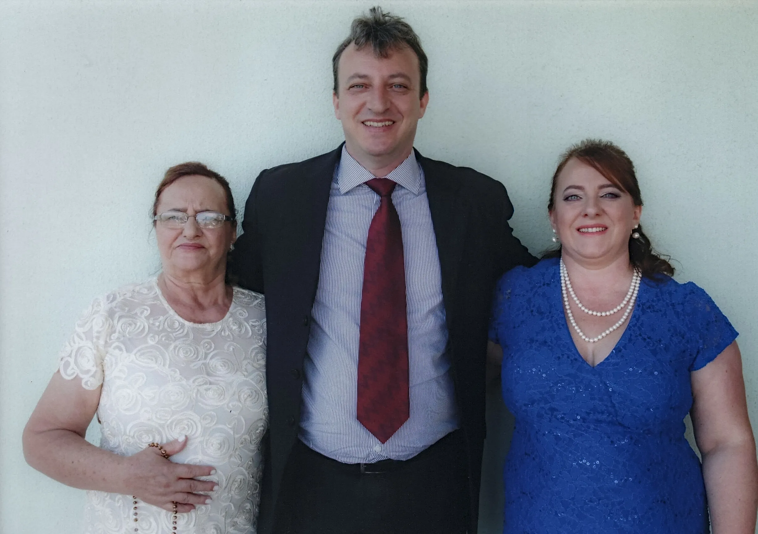 Um homem sorridente de terno listrado em azul e branco, com uma gravata vermelha listrada, está entre duas mulheres. A mulher da esquerda é uma senhora de óculos e cabelo ruivo encaracolado, vestindo uma blusa branca texturizada e segurando um rosário. A mulher da direita, com cabelo ruivo e um colar de pérolas, veste um vestido azul texturizado.