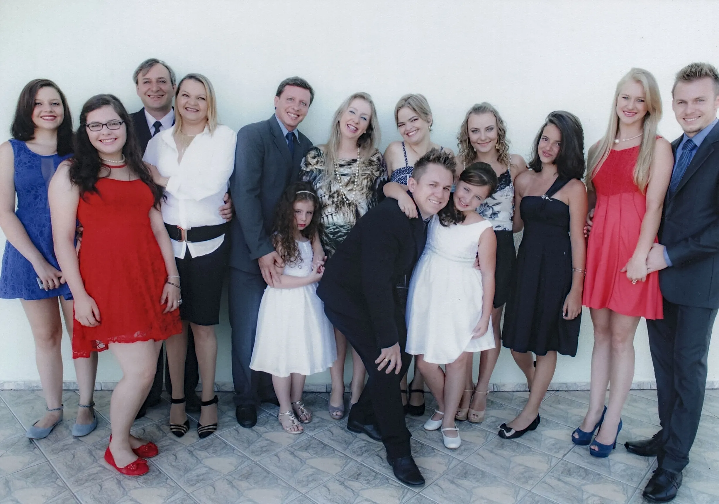 Um grupo de quinze pessoas, incluindo três crianças, posa informalmente para uma foto contra uma parede branca. Eles estão vestidos elegantemente em trajes de festa e sorriem para a câmera, com muitos casais entrelaçados ou próximos, e os homens usam ternos escuros.