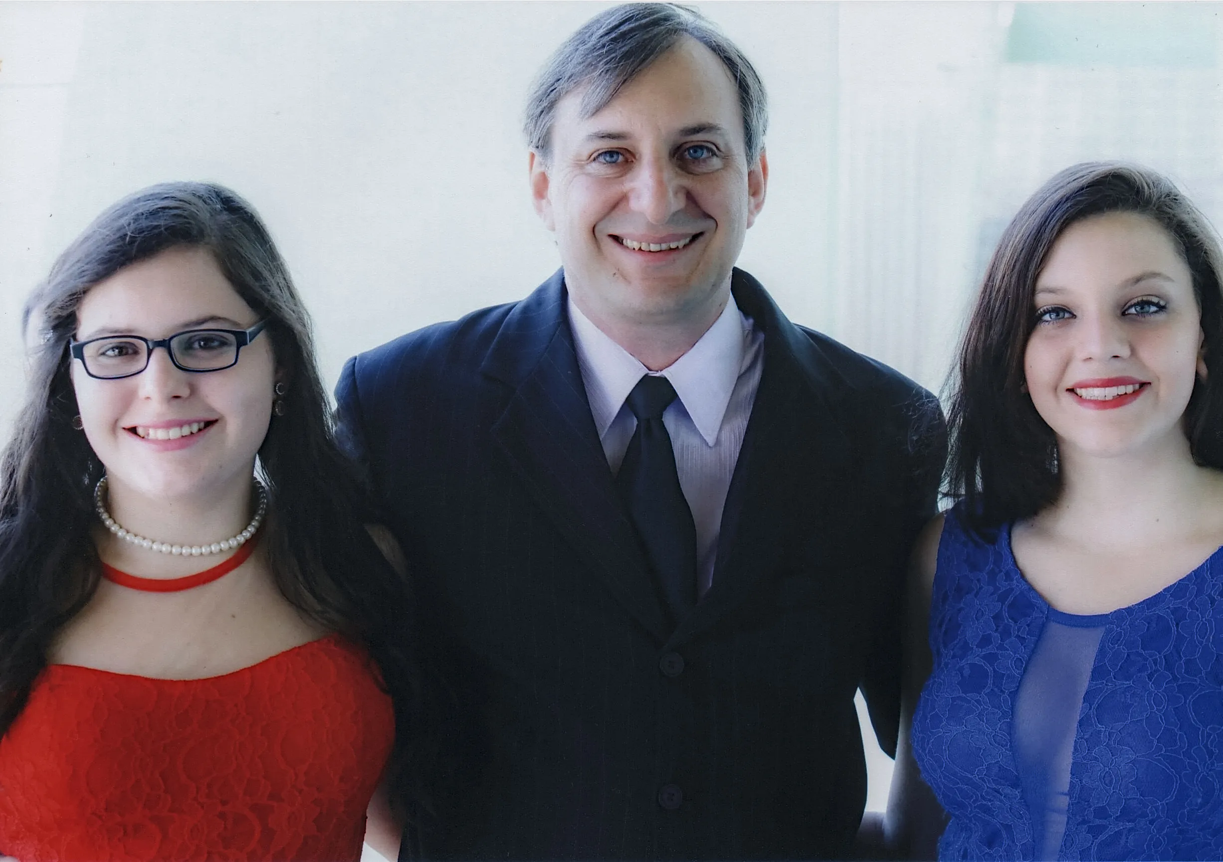 Homem de terno escuro sorrindo entre duas mulheres. A da esquerda usa um vestido vermelho e óculos, enquanto a da direita usa um vestido azul.
