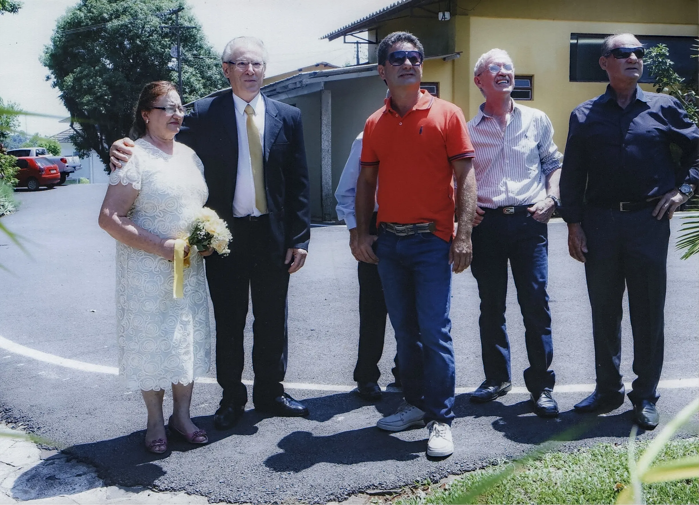 Um grupo de cinco pessoas está posando numa rua. Um homem e uma mulher, em primeiro plano, abraçam-se, a mulher segurando um buquê.